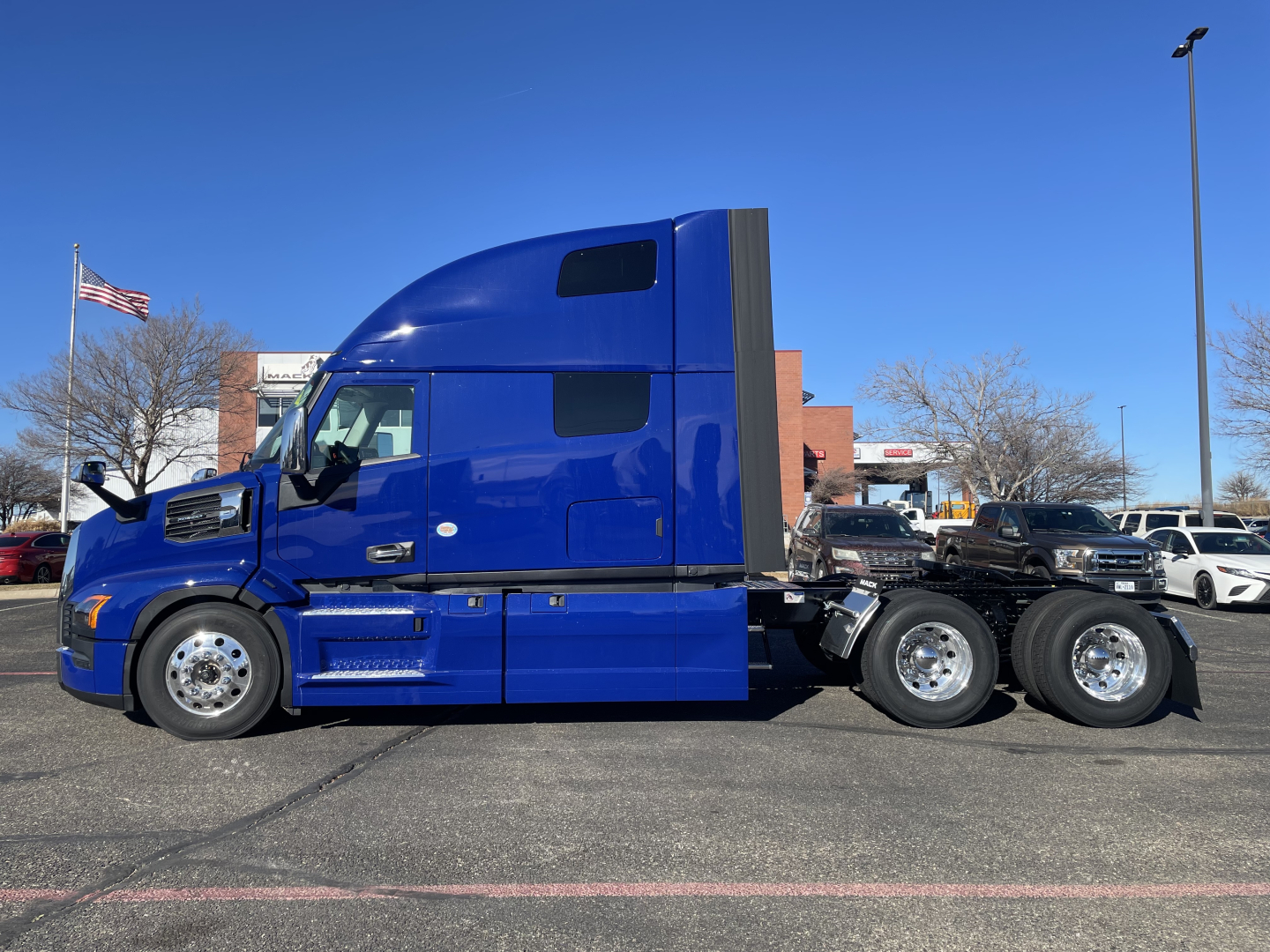 Truck side profile showing blue Mack PR64T76H sleeper cab with three rear axles and chrome wheels.