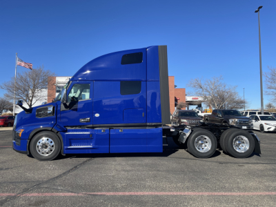 Truck side profile showing blue Mack PR64T76H sleeper cab with three rear axles and chrome wheels.