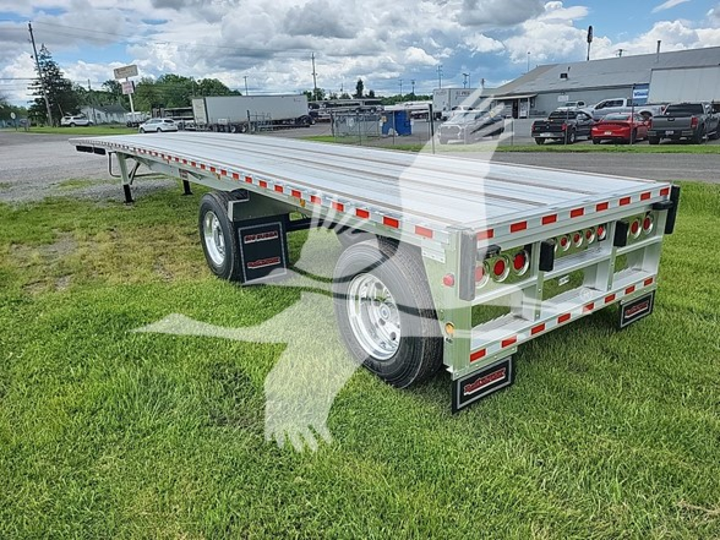 Flatbed trailer with aluminum deck and rear ramps showing reflective tape and multiple tail lights, single axle and dual wheels, set on grass at a dealership lot.