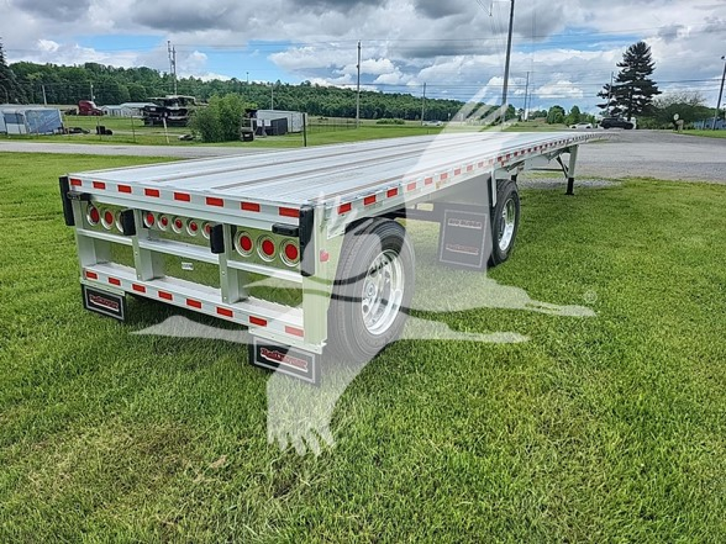 Flatbed trailer in an outdoor setting showing rear and left-side with multiple round taillights, reflective tape, and dual wheels; model tag and TR065705 visible.