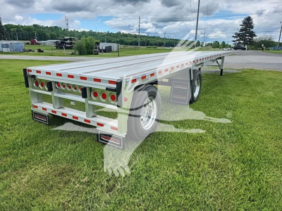Flatbed trailer in an outdoor setting showing rear and left-side with multiple round taillights, reflective tape, and dual wheels; model tag and TR065705 visible.