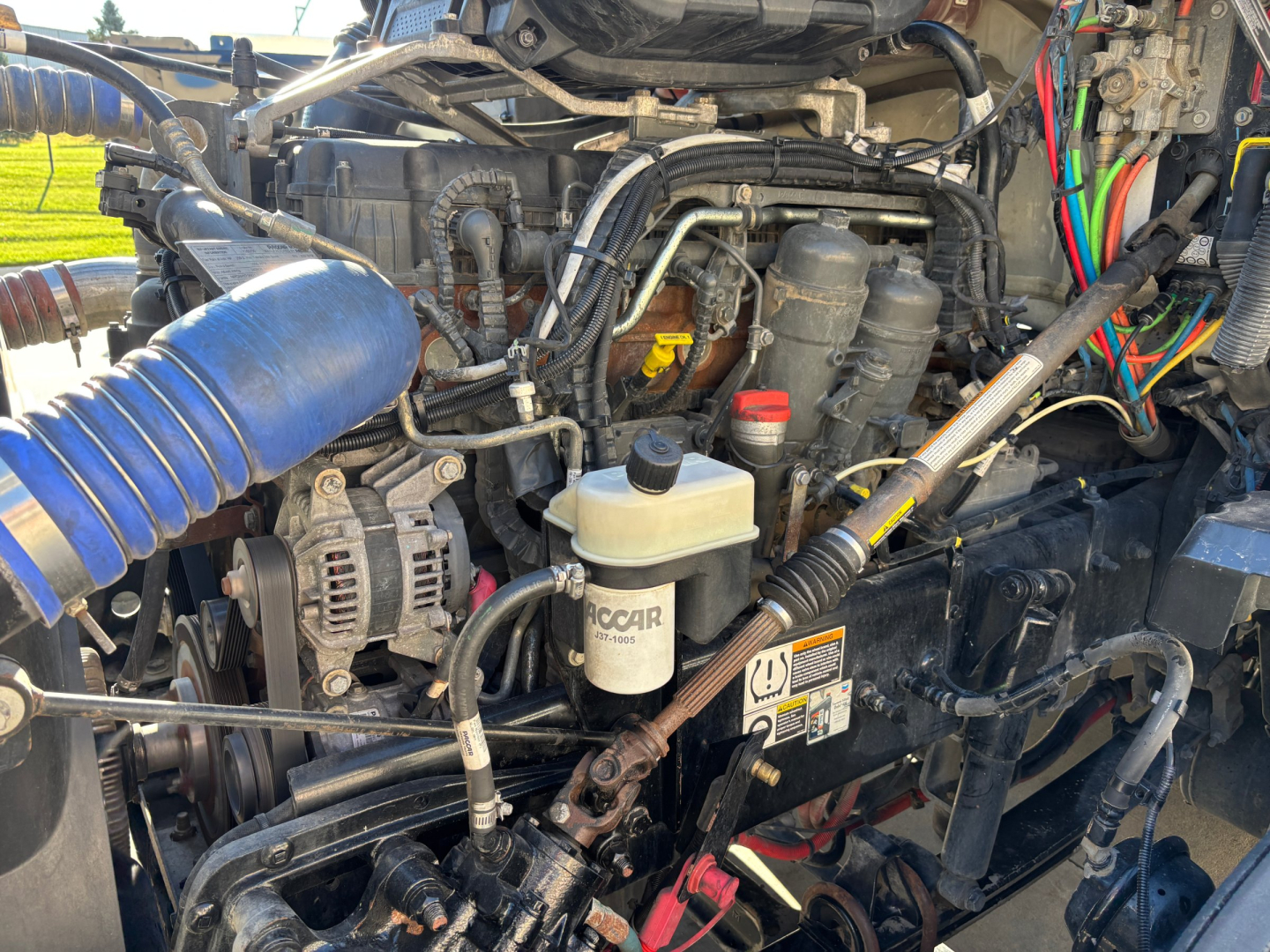 Kenworth T680 engine bay with visible hoses, reservoir, and wiring harnesses; components include coolant reservoir, air lines, and electrical connectors.