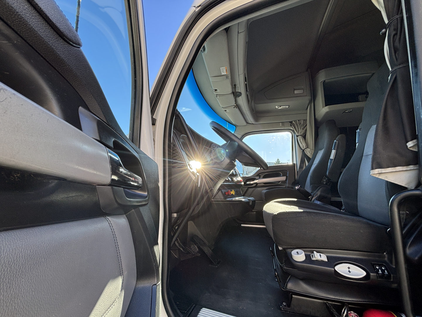 Interior of a Kenworth T680 cab showing driver seat controls, steering wheel, and dashboard from the open driver door.