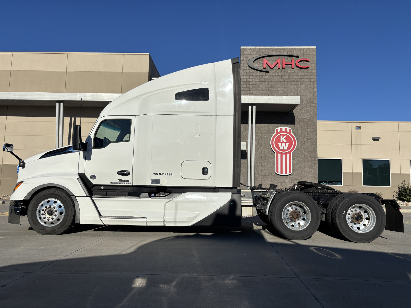 Kenworth T680 tractor unit side view showing cab and rear wheels; dealership building in background with MHC logo.