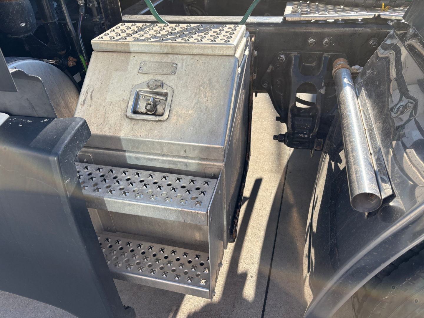 Kenworth truck engine bay area showing a metal toolbox with vented steps and a small latch, adjacent to cylindrical exhaust or exhaust pipe and chassis components.
