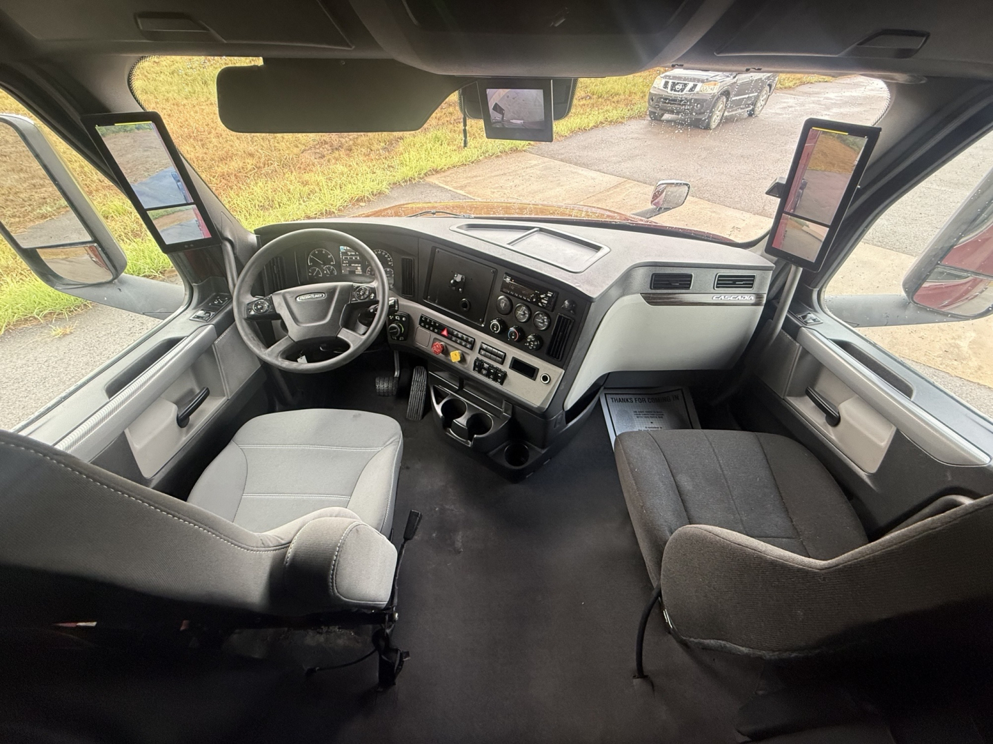 Truck cab interior showing steering wheel, dashboard, dual large screens on mirrors, driver and passenger seats, center console with controls.