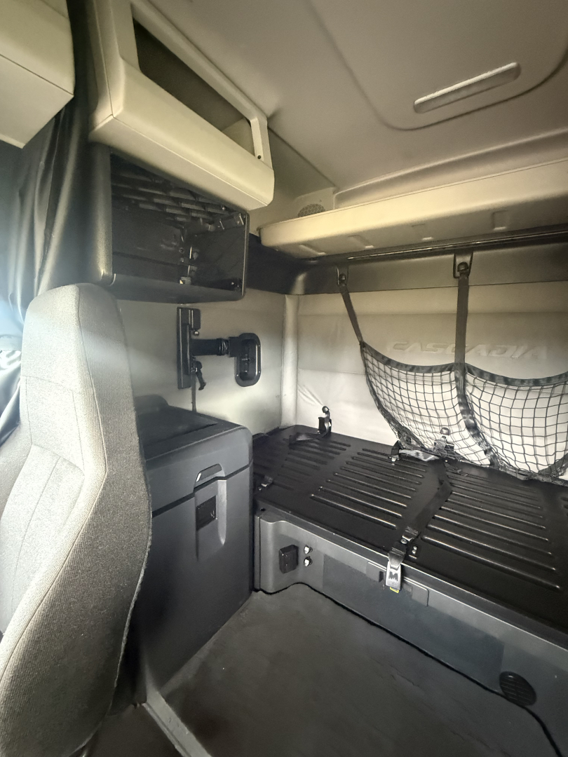 Interior sleeper cab cargo area of a Freightliner Cascadia 126 showing lower storage area, binders, and side ladder rail with netted pocket.