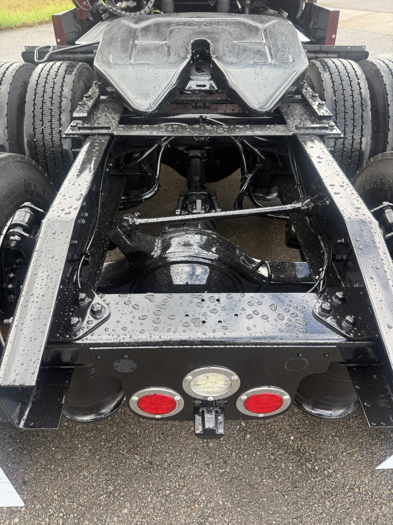 Truck chassis rear view showing suspension, drive axles, and fifth wheel area with wet surface and multiple red/white reflectors.