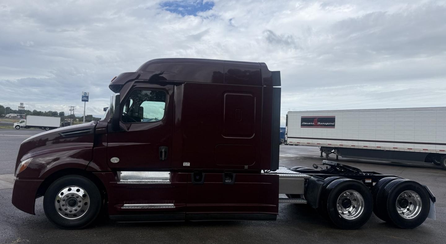 Freightliner Cascadia 126 tractor cab, side profile view showing maroon exterior, sleeper cab, dual rear axles, chrome fuel tanks, and trailer drop-frame connection.