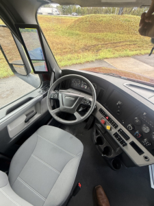 Truck cab interior showing steering wheel, dashboard with gauges, and driver seat in a Freightliner Cascadia cabin.