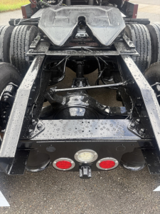 Truck chassis rear view showing suspension, drive axles, and fifth wheel area with wet surface and multiple red/white reflectors.