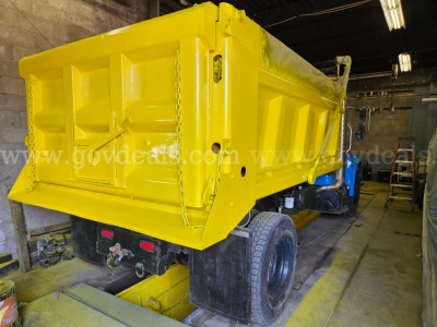 Dump truck bed of a yellow 1999 International 4700 dump truck in a shop, rear view showing tailgate, hydraulic lines, and dual rear wheels.