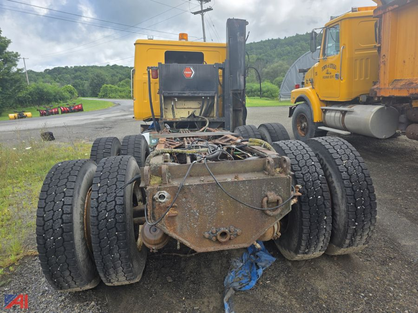 Rear view of a Volvo WG cab and chassis showing the back of the frame and multiple rear wheel assemblies; rusted drivetrain area; two yellow trucks in background.