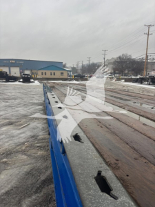 Traveling axle trailer bed with wooden deck and blue side railing, showing wear along the deck, from a low-angle outdoor view.
