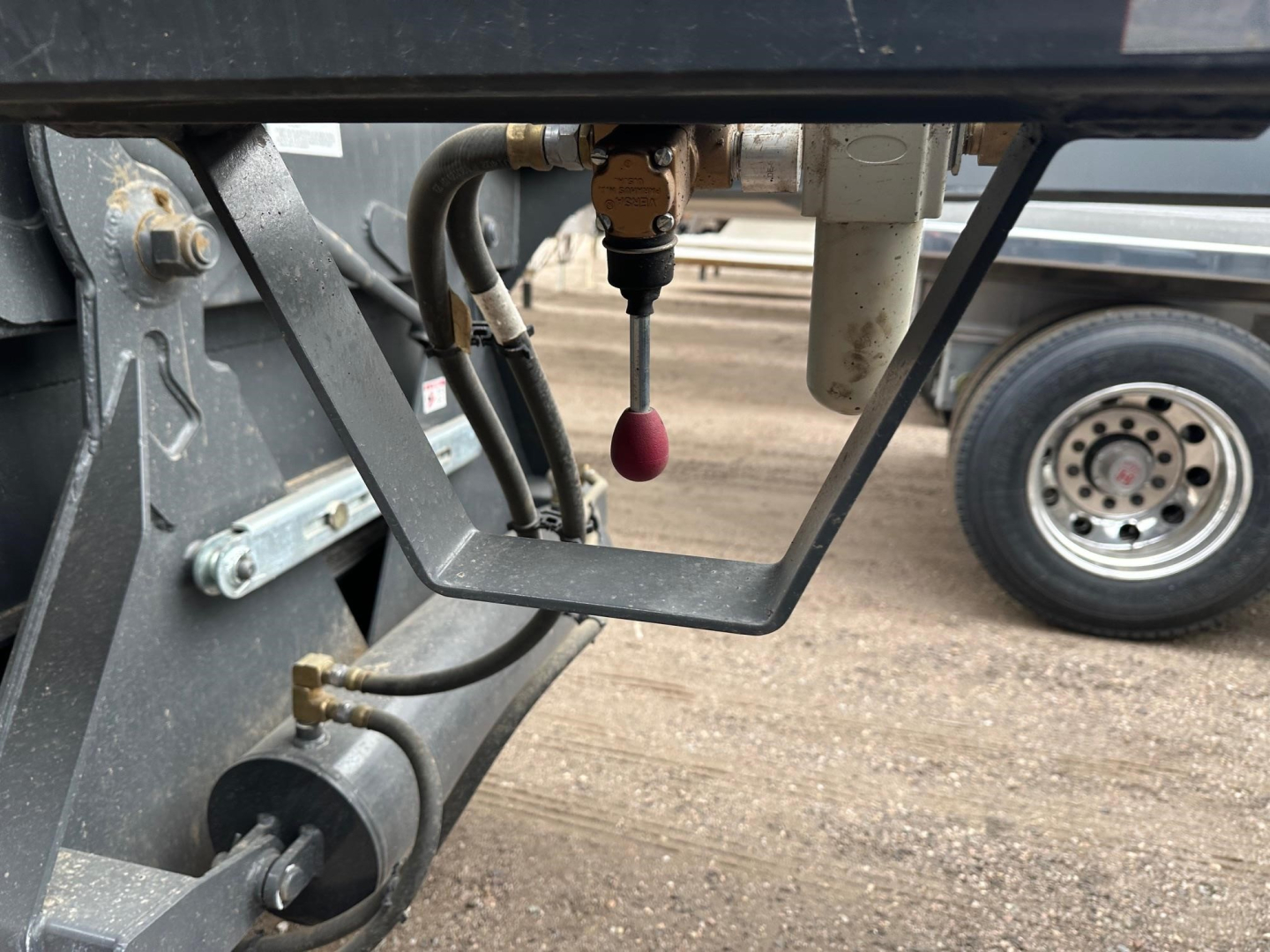 Trailer bottom dump connection with hydraulic lines and control valve, red round pull handle, metal frame, wheel in background.