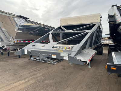 Bottom dump trailer frame showing steel structure with level indicators and platform, gray color, tilted hopper at rear, side ramps and hitching area visible.