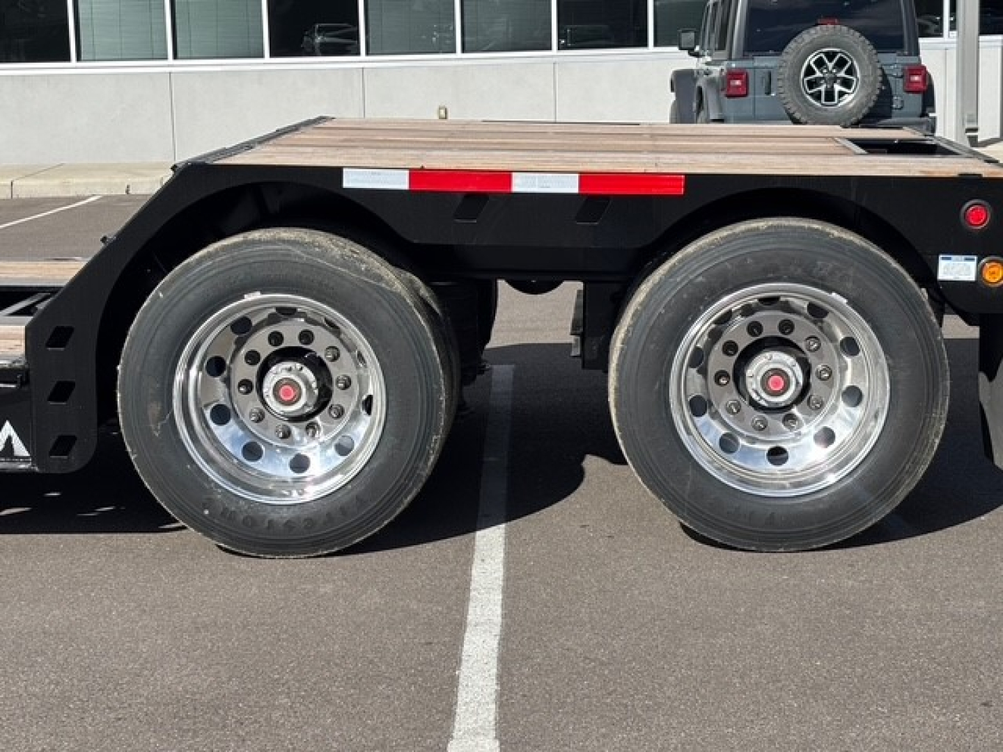 Dual rear axles of a lowboy trailer with polished aluminum wheels and wooden deck, side of a truck in background.