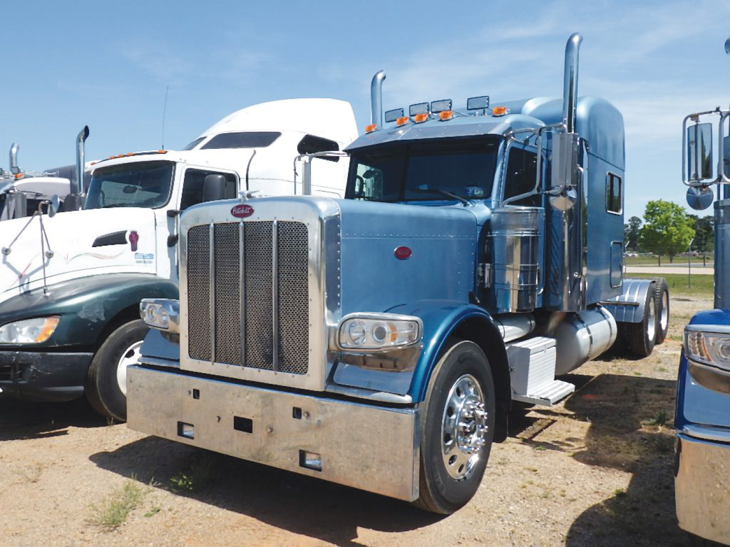Peterbilt 389 UltraCab Extended Hood tractor in blue, front view showing large chrome bumper, vertical grille, dual vertical exhaust stacks, chrome wheels, and dual rear wheels; other trucks in background.