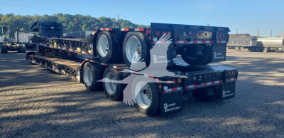 Lowboy trailer showing tandem axle suspension and long flatbed with dual deck; branding on mudflaps reads The American…; VIN sticker on front area not visible.