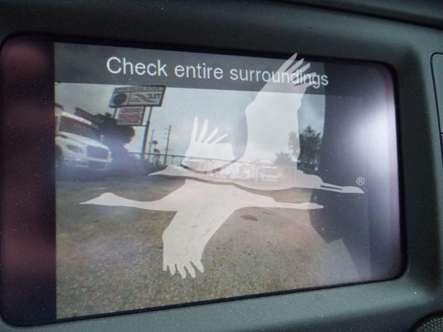 Vehicle display screen showing a warning: 'Check entire surroundings' with reflection of a logo and a bird graphic.