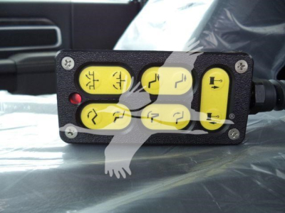 Winch control pendant showing six yellow push buttons with black icons, mounted on a black rectangular housing with screws at corners, red indicator light, wired to truck equipment.