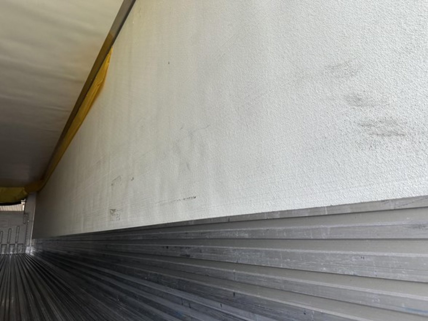 Trailer side shown with white upper wall and a ribbed metal lower edge; yellow curtain or tarp at top left. No legible model text visible.