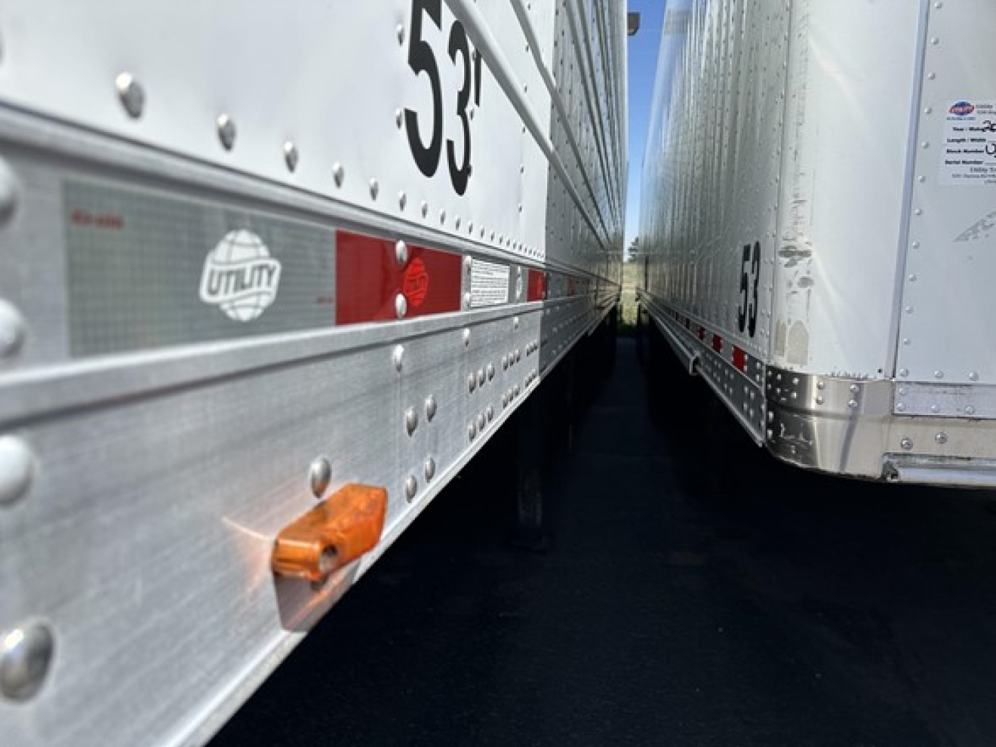 Trailer side view showing Utility brand decal and reflective strips between two refrigerated trailers, close-up midsection with riveted aluminum panels.
