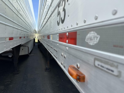 Two long utility trailers side by side with riveted aluminum sides, showing '53' and reflections; orange side marker light and small label sticker visible on the right trailer