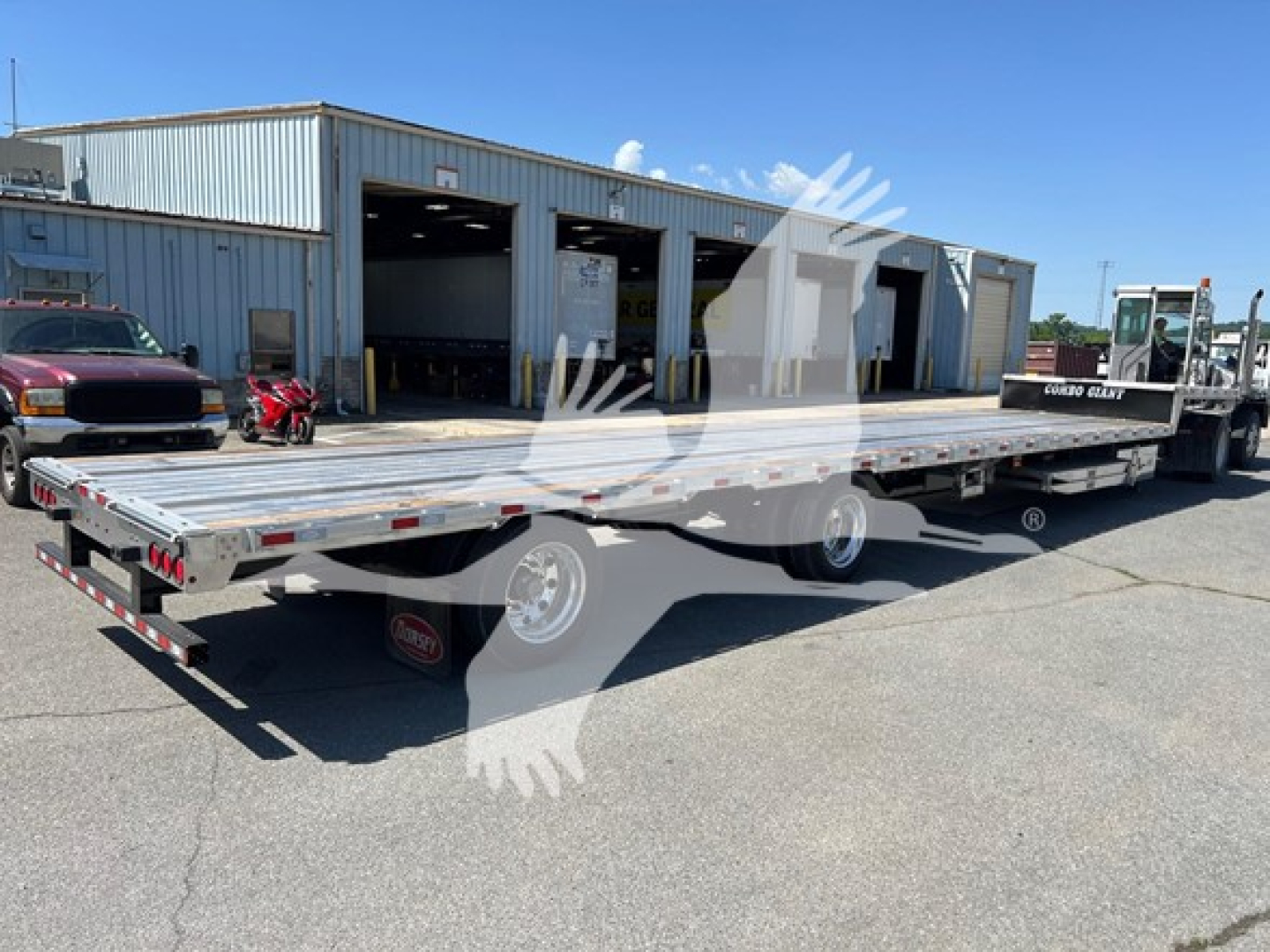 Trailer showing a long flatbed with ramp kit and multiple axles in a lot; site name and loading bays in background.