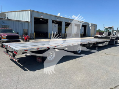 Trailer showing a long flatbed with ramp kit and multiple axles in a lot; site name and loading bays in background.