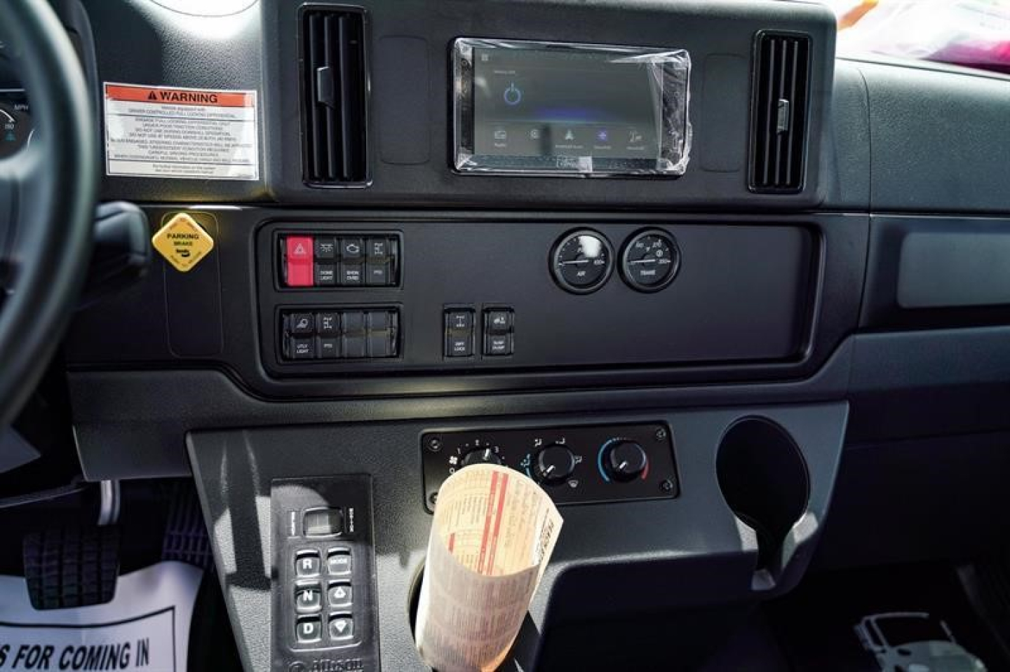 Cab interior panel of a Freightliner Business Class M2 showing dashboard controls, steering wheel, and center console with switches, gauges, and a wrapped label on the gear area.
