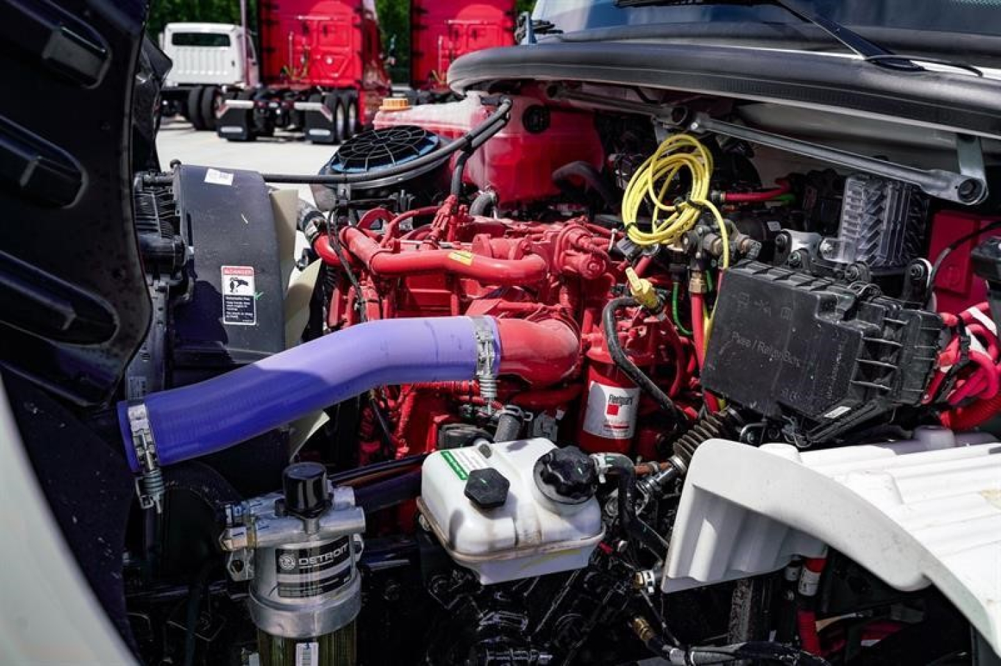 Freightliner M2 106 engine bay showing red engine, purple coolant hose, yellow cables, and various hoses and reservoirs.
