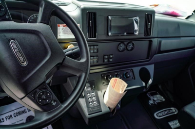 Frontal interior view of a Freightliner Business Class M2 106 Plus cab dashboard and steering wheel with controls and a paper tag.