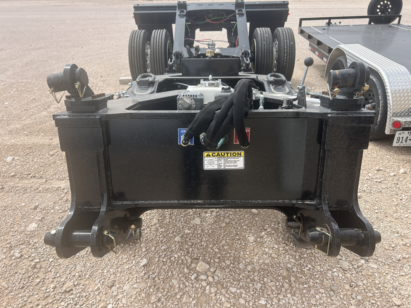 Flatbed trailer hitch assembly showing a black metal hitch frame with coiled cables and hydraulic components, caution sticker, and two bottom hooks.