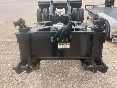Flatbed trailer hitch assembly showing a black metal hitch frame with coiled cables and hydraulic components, caution sticker, and two bottom hooks.