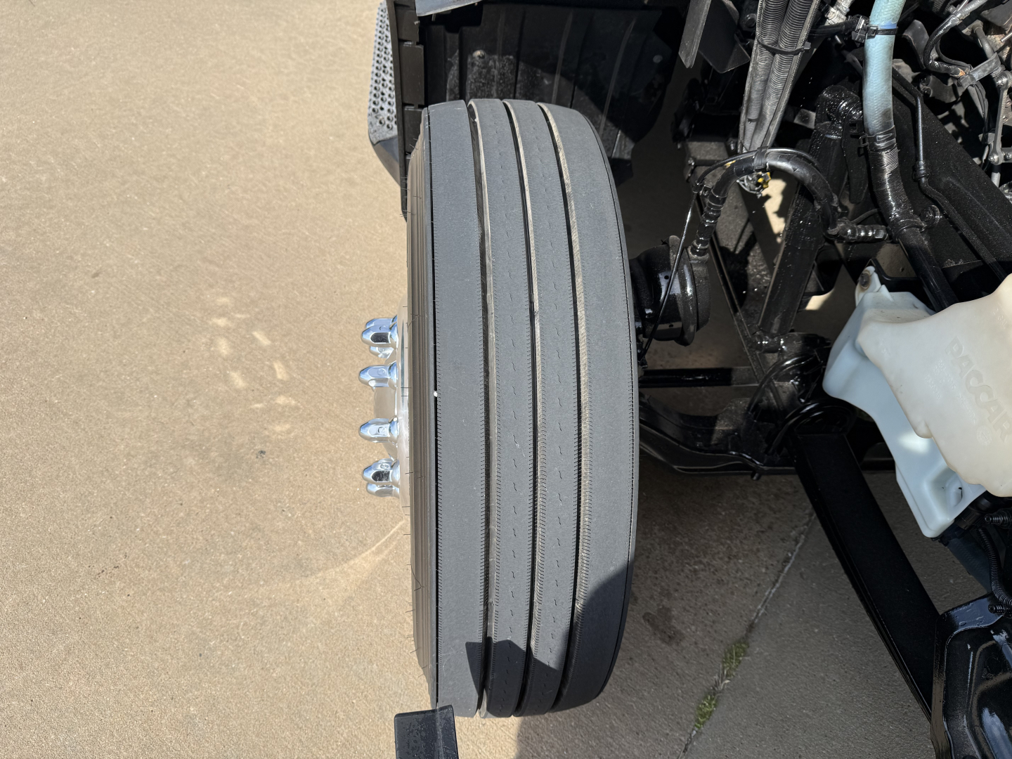 Rear view of a 2023 Peterbilt 579 front drive axle showing multiple hub and dual tires vertical stack with wiring, white coolant reservoir on right, and utility components.
