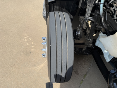 Rear view of a 2023 Peterbilt 579 front drive axle showing multiple hub and dual tires vertical stack with wiring, white coolant reservoir on right, and utility components.
