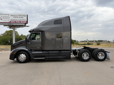 Peterbilt 579 semi-truck tractor in-gray cabin profile with sleeper cab and tandem rear wheels, side window, fuel tank, and a small billboard in background.