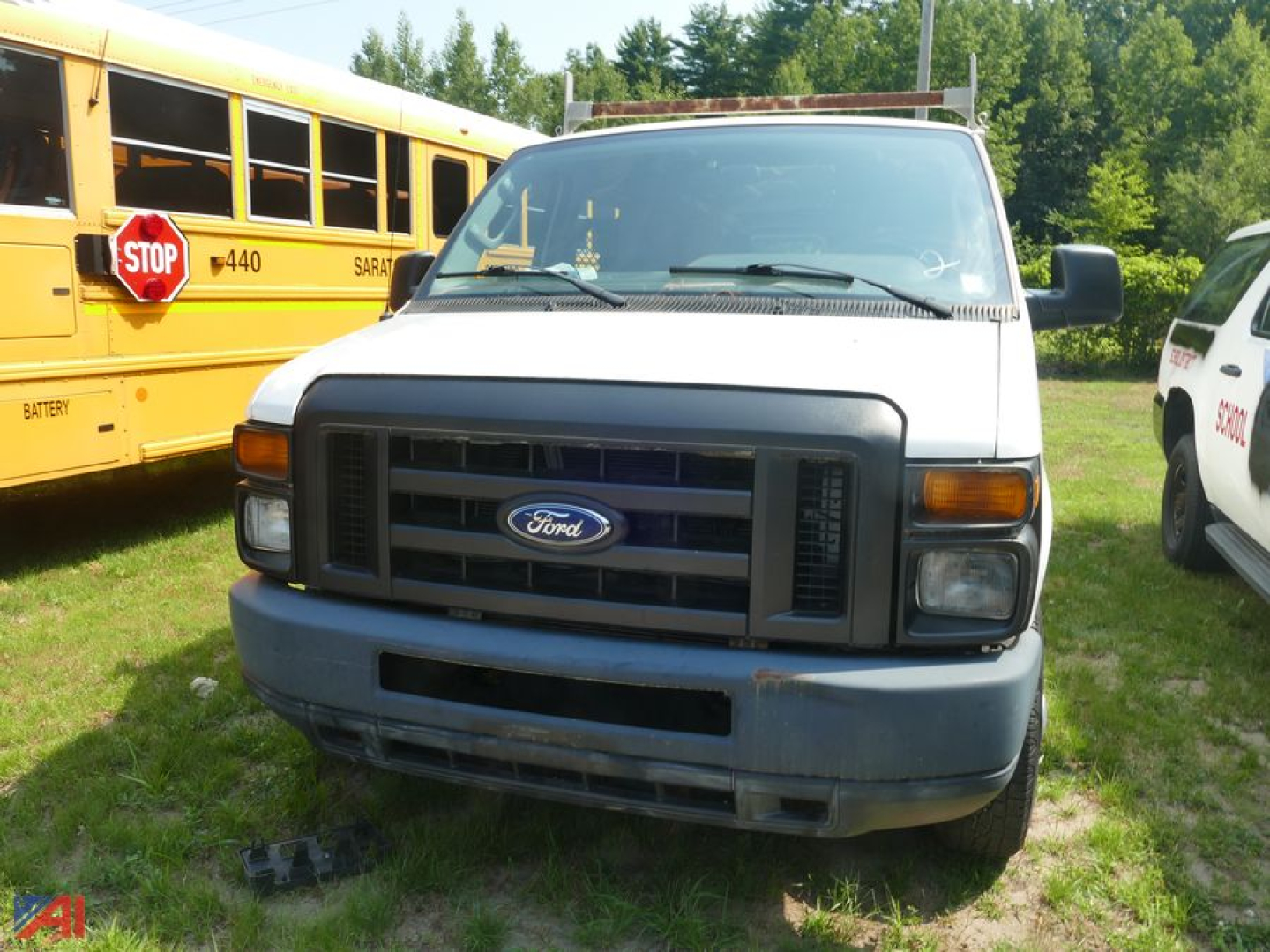 Front view of a white 2014 Ford E150 full-size cargo van with black grille, Ford emblem, and blue bumper in a grassy lot, other vehicles nearby.
