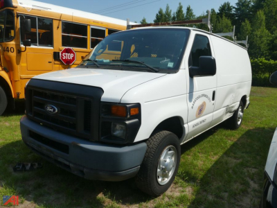 Ford E-150 cargo van in white, front-left view, side logo visible, parked on grass with a yellow school bus in background.