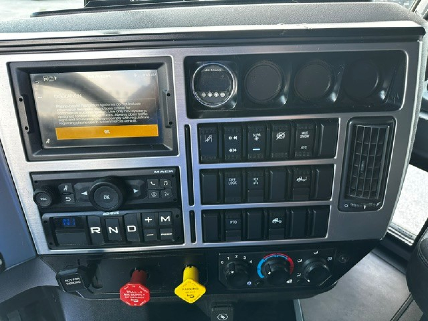 Mack AN64T70 truck dashboard with touchscreen display, various switches, gauges, and climate controls; gear selector shows R N D plus M; emergency/disconnect buttons visible.
