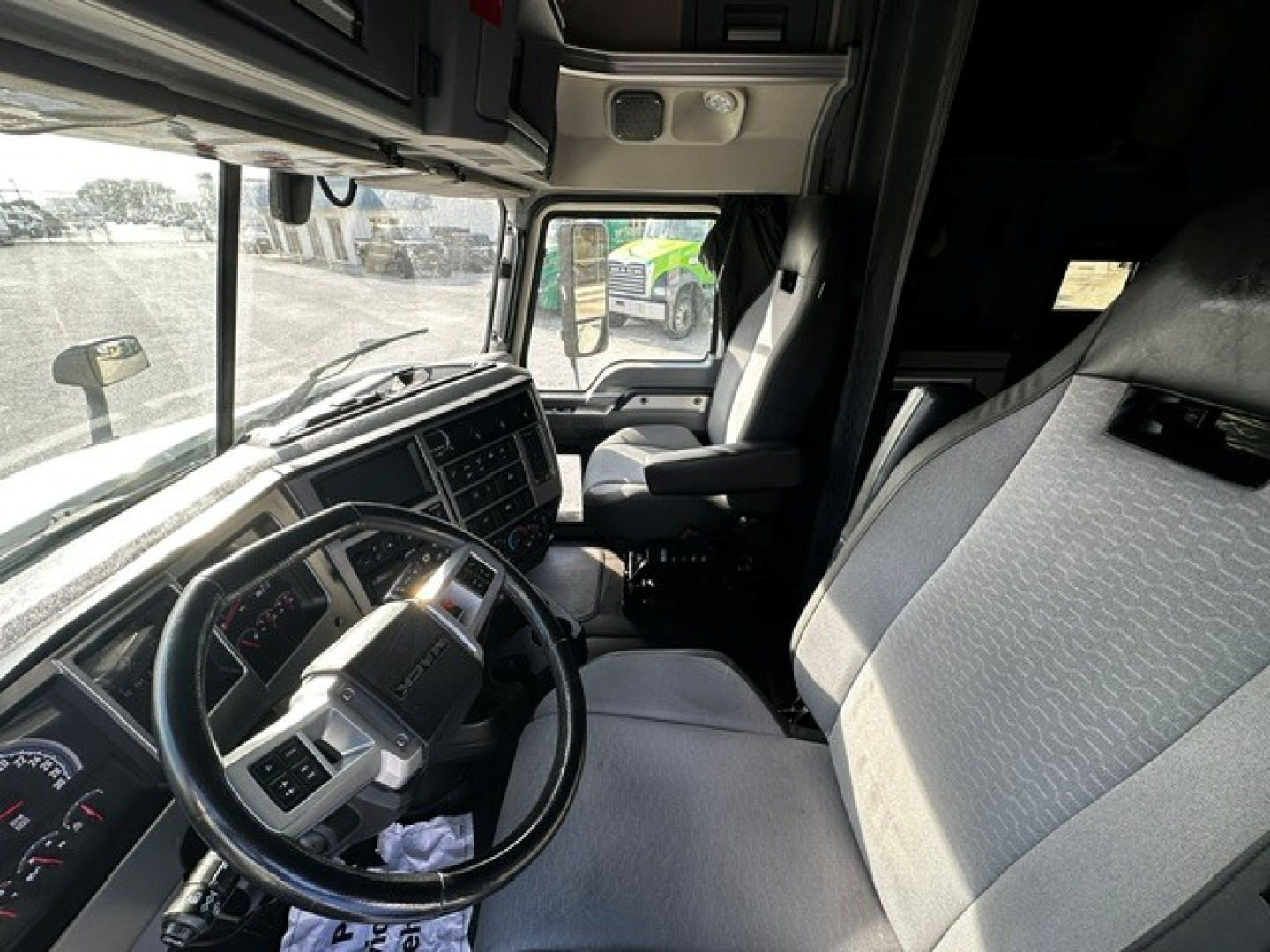 Truck cab interior of a 2020 Mack AN64T70 show dashboard, steering wheel, driver's seat with light gray/black upholstery; view toward passenger area and windshield