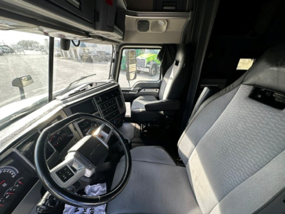 Truck cab interior of a 2020 Mack AN64T70 show dashboard, steering wheel, driver's seat with light gray/black upholstery; view toward passenger area and windshield