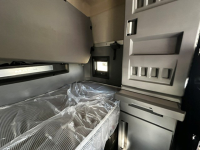 Mack AN64T70 truck sleeper area interior with plastic-wrapped seat/bed in foreground and cabinetry panels; window opening visible.