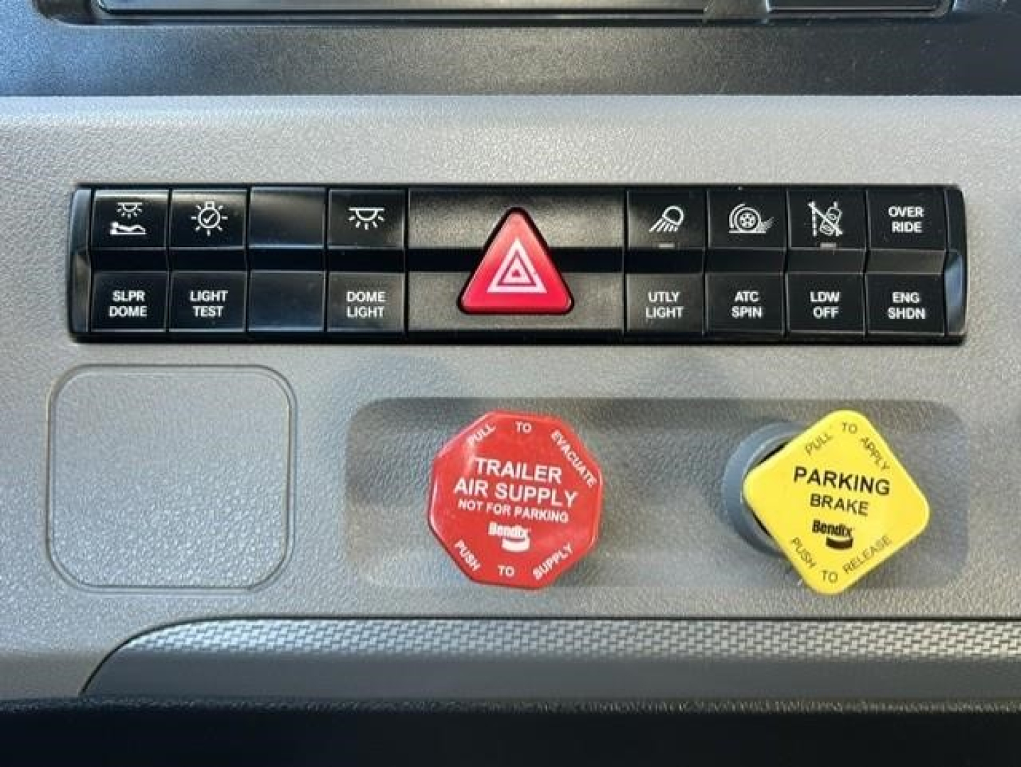 Dashboard switches and knobs in a Freightliner Cascadia showing hazard light button, parking brake and air supply handles, and various indicator symbols.