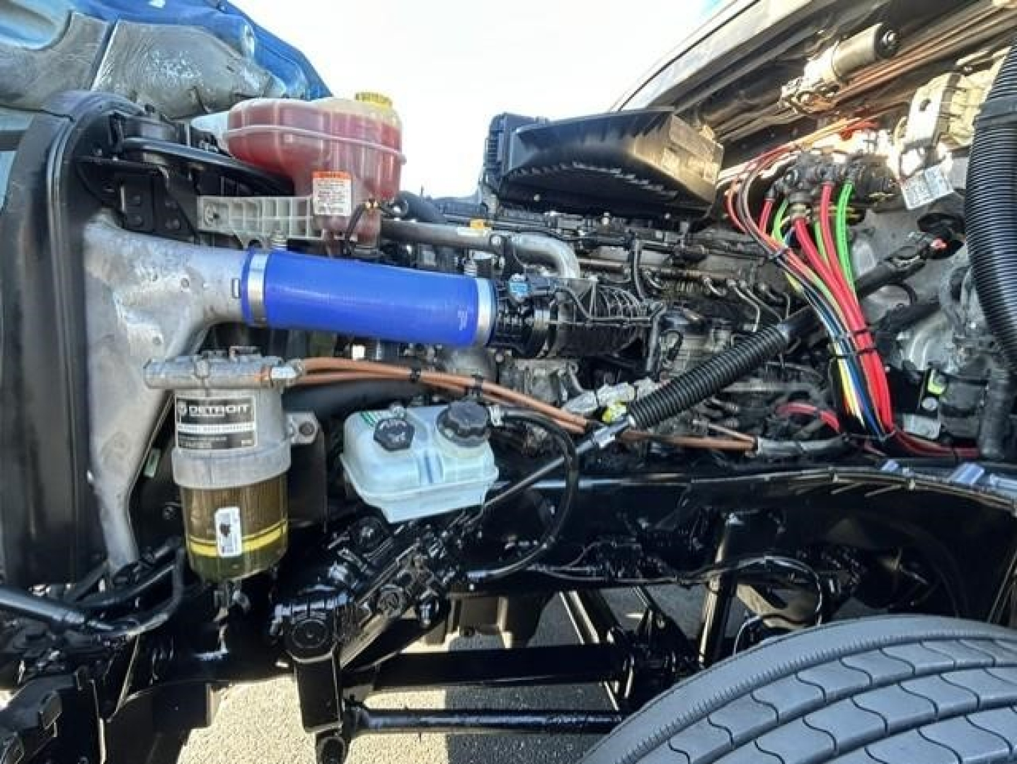 Conventional sleeper truck engine bay of a 2022 Freightliner Cascadia showing blue intercooler hose, fuel filter, coolant reservoir, wiring harness, and various hoses.