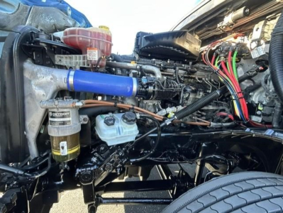 Conventional sleeper truck engine bay of a 2022 Freightliner Cascadia showing blue intercooler hose, fuel filter, coolant reservoir, wiring harness, and various hoses.