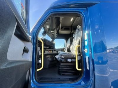 Interior of a 2022 Freightliner Cascadia cab with blue exterior; driver door open showing black seats with plastic coverings, yellow grab handles, dashboard and steering wheel.