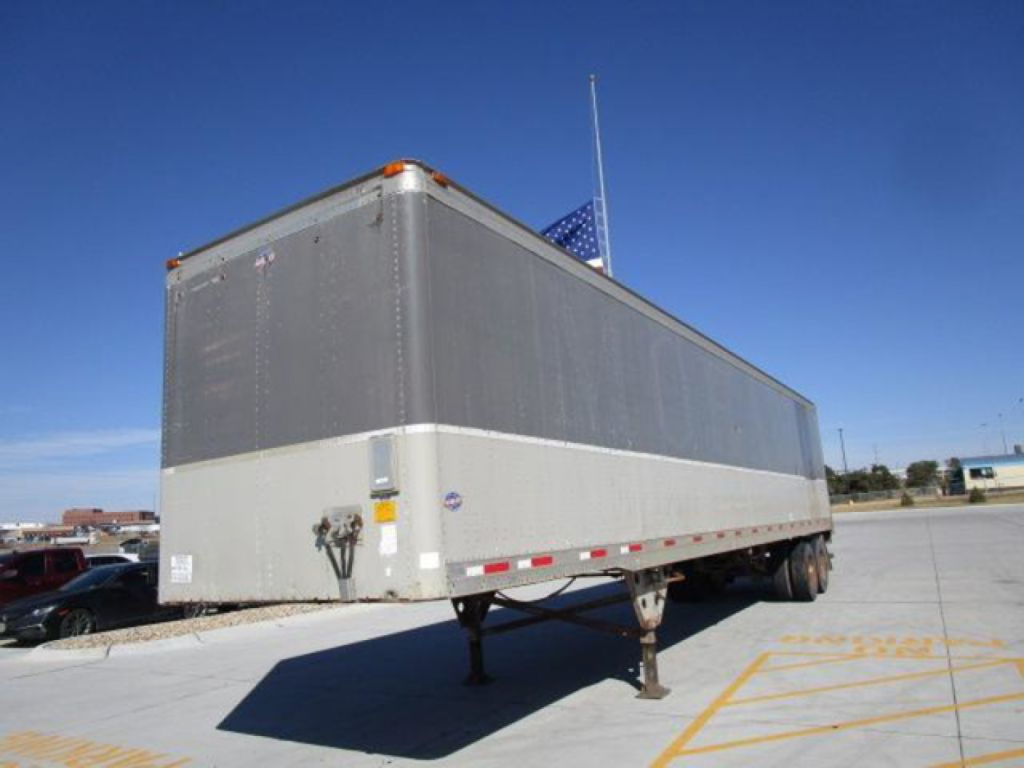 Utility trailer/van on trailer legs in a parking lot, with silver upper and light gray lower body, long rectangular box, dual rear wheels visible, blue sky.
