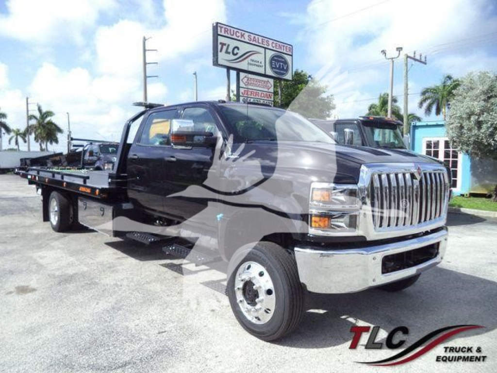 Truck with flatbed tow body in a dealership lot, International CV cargo/tow truck visible with chrome front grille and large side mirrors.